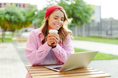 Süslü kırmızı şapkalı genç bir kadının portresi, elinde kahve bardağı, sokakta oturuyor, dizüstü bilgisayar kullanıyor, web sitesi, sipariş veriyor. Çevrimiçi alışveriş kavramı