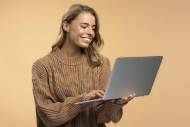 Güzel, gülümseyen bir kadının portresi. Dizüstü bilgisayar kullanıyor. Kahverengi süveter giyiyor ve bej arka planda klavyeye yazıyor. Teknoloji konsepti