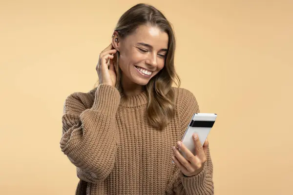Smiling modern woman wearing stylish brown sweater holding mobile phone, using website, video call isolated on beige background. Communication concept
