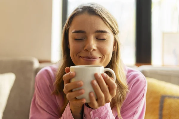 Closeup portrait of attractive smiling woman with eyes closed holding cup of aroma coffee, enjoying hot drink sitting in modern cafe. Coffee break concept