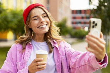 Kırmızı şık şapkalı çekici bir kadın elinde kahve fincanı ve akıllı telefonuyla sokakta selfie çekiyor. Mobil uygulama kayıt videosu kullanan etkili, parkta iletişim kurabilen 
