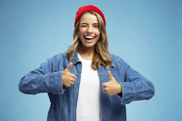 Cute smiling beautiful woman winking wearing stylish yellow hat, jeans jacket holding thumb up isolated on background. Portrait of happy modern hipster female looking at camera. Positive lifestyle