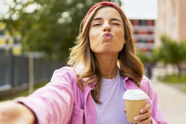 Funny face woman wearing red stylish hat holding cup of coffee taking selfie on the street. Happy successful blogger influencer recording video, communication online outdoors. Technology