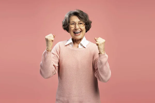 Portrait smiling funny senior woman, happy modern grandmother holding hands win something celebration success isolated on pink background. Good news, positive lifestyle concept
