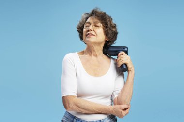 Mutlu son sınıf kadını, modern büyükanne masaj tabancası kullanıyor mavi arka planda kendine masaj yapıyor. 