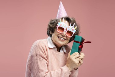 Komik gözlükler takan, gülümseyen ve pembe arka planda izole edilmiş doğum günü kutlaması yapan son sınıf öğrencisi bir kadın. Tatil partisi konsepti 
