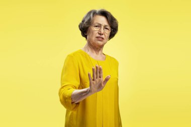 Sarı arka planda izole edilmiş kameraya bakarken dur işareti gösteren ciddi bir gözlük takan yaşlı kadın portresi. Hareketli kavram