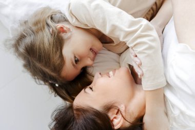 Çekici, gülümseyen bir anne ve küçük kızın beyaz yatak çarşafları üzerinde uzanmış, birbirlerine sarılmış portresi. Annelik kavramı, reklam.