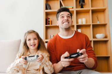 Gülümseyen, yakışıklı bir adam ve kızının video oyunları oynayıp, elinde joystick tutarak rahat bir odada oturduğu portre. Çevrimiçi teknoloji kavramı