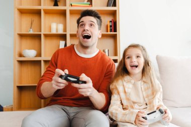 Heyecanlı küçük baba ve küçük kızın portresi oyun konsolu tutuyor, video oyunları oynuyor, evde rahat bir odada oturuyor, eğleniyor. Çevrimiçi teknoloji kavramı