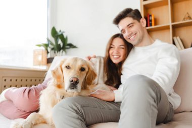 Çekici genç bir çiftin portresi, çekici erkek ve kadın Golden retriever 'ı okşayarak rahat bir kanepede otururken ev seçiciliğine odaklanıyor. Aşk kavramı, hayvan bakımı