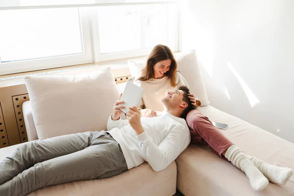 Portrait of young family, handsome man holding book, smiling, lying on comfortable rug next to beautiful woman, relaxing together in cozy home. Relationship concept