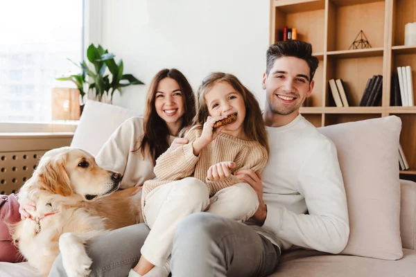 Beautiful smiling family, happy mother, father and cute daughter eating cookies, looking at camera, sitting on comfortable sofa with pet golden retriever. Concept of parenthood, love