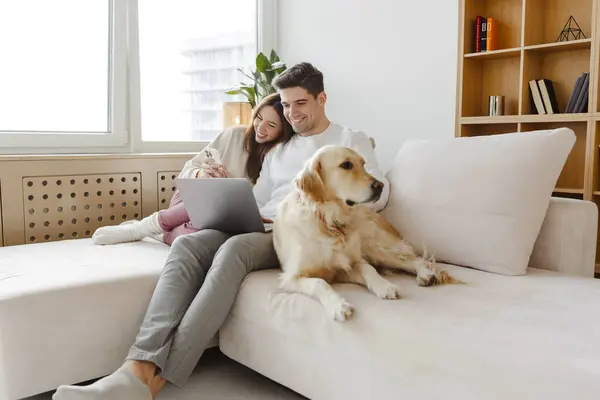 Portrait of happy young couple using laptop, man holding mobile phone, hugging dog, sitting together on comfortable sofa at home. Online shopping concept