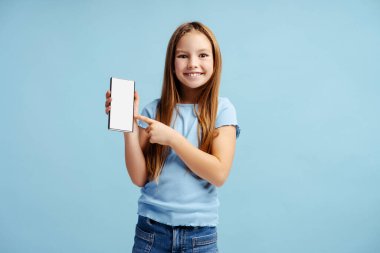 Sıradan kıyafetler giyen, cep telefonu tutan ve mavi arka planda izole bir şekilde duran küçük mutlu kızın portresi. Teknoloji konsepti 