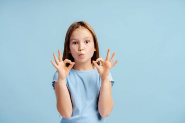 Moda tarzı küçük bir kızın portresi, günlük tişört giyen, kameraya bakan, mavi arka planda izole edilmiş güzel hareketlerle poz veren bir model. Reklam kavramı
