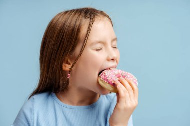 Mavi arka planda tek başına dururken pembe çörek yiyen mutlu küçük kızın portresi. Yemek kavramı, tatlı. 