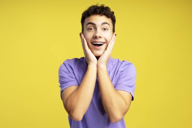 Çekici heyecanlı genç çocuğun portresi, diş telleri ile kameraya bakıyor, şık mor tişört giyiyor ve sarı arka planda tek başına duruyor. Alışveriş konsepti