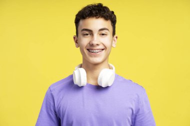 Diş teli takmış, kulaklık takmış, müzik dinleyen, sarı arka planda izole edilmiş kameraya bakan mutlu ve gülümseyen çocuğun portresi. Teknoloji konsepti