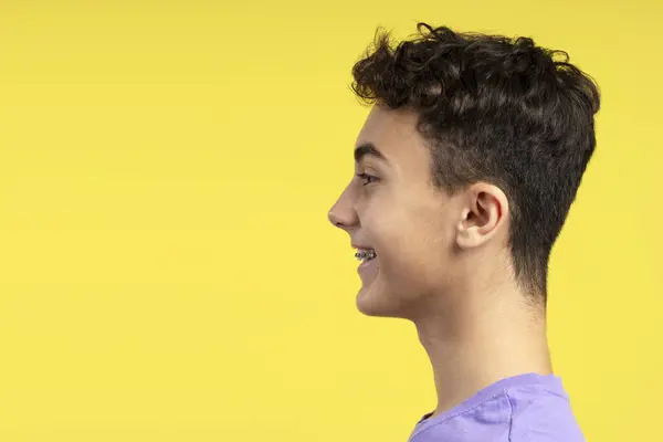 Side view portrait of handsome smiling young boy with dental braces looking away standing isolated on yellow background copy space. Treatment concept, advertisement