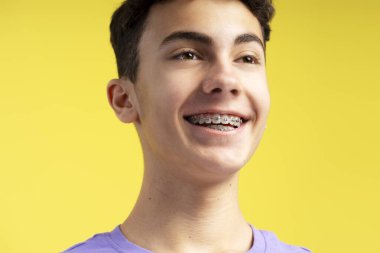 Dişlerinde diş teli olan gülümseyen, kendine güvenen genç bir çocuğun sarı arka planda izole edilmiş bir şekilde bakışları. Sağlık hizmeti, ortodontik konsept 