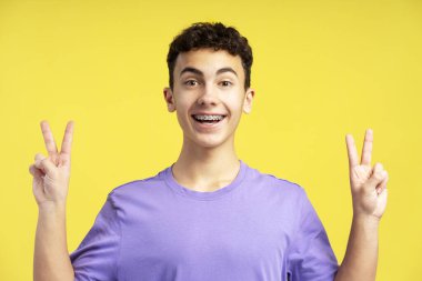 Dişlerinde diş teli olan gülümseyen genç çocuk sarı arka planda izole edilmiş kameraya bakarak zafer işareti gösteriyor. Pozitif duygular, sağlık hizmetleri, ortodontik kavramlar