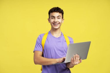 Sırt çantalı gülümseyen genç çocuk sarı arka planda dizüstü bilgisayar tutuyor. Çevrimiçi eğitim kavramı 