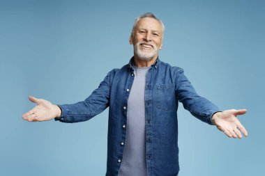 Yakışıklı, gülümseyen, gri saçlı, şık, sıradan bir kot gömlek giyen, mavi arka planda izole edilmiş kolları açık kameraya bakan bir adamın portresi. Pozitif yaşam tarzı kavramı