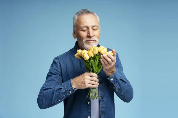 一个面带微笑的英俊老人的画像 他穿着时髦的斜纹棉布衬衫 拿着一束花 黄色的郁金香 紧闭的眼睛将它们隔离在蓝色的背景中 广告概念 — 图库照片
