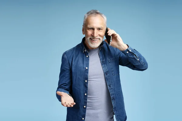 一个面带微笑 迷人而成熟的60岁男人拿着手机看着摄像机的画像 一边接电话 一边说着一些孤立的蓝色背景的话 传播概念 — 图库照片