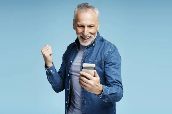 面带微笑的灰白头发的成熟男子手持手机 观看视频 在蓝色背景下孤立地做出胜利姿态 高级男性检查邮件 技术概念 — 图库照片