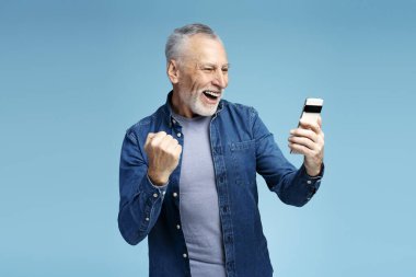 Gülümseyen emeklilerin portresi, elinde cep telefonu olan olgun bir adam, mavi arka planda zafer jesti yapan video izliyor. Son sınıf erkek postaları kontrol ediyor. Teknoloji konsepti