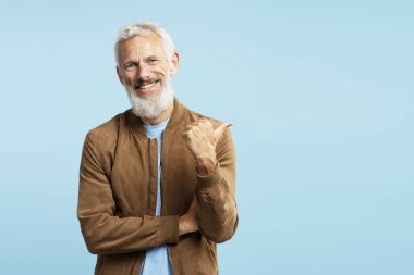 Gülümseyen, gri saçlı yakışıklı bir adamın portresi, mavi arka planda izole edilmiş şık kahverengi ceket giyen sakallı bir hippi, fotokopi alanını işaret ediyor. Alışveriş, mağaza konsepti