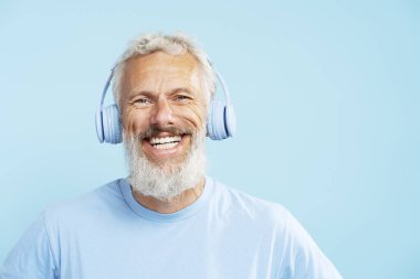 Mutlu, olgun bir adam, gri saçlı hippi mavi arka planda izole edilmiş kablosuz kulaklıkla müzik dinliyor. Pozitif yaşam tarzı, teknoloji kavramı