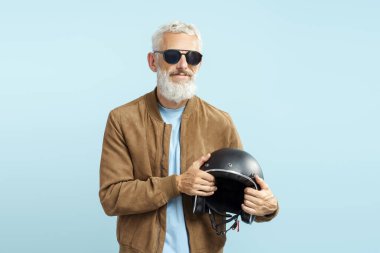 Yakışıklı, olgun bir adam, gri saçlı, sakallı, şık güneş gözlüğü takan, kahverengi deri ceketli, mavi arka planda izole edilmiş motosiklet kaskı tutan.