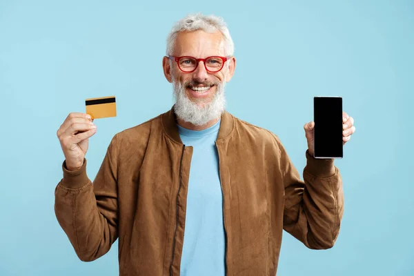 Smiling confident mature man, bearded hipster holding golden credit card, mobile phone shopping online, showing smartphone screen isolated on blue background. Sale, cash back, bank client concept