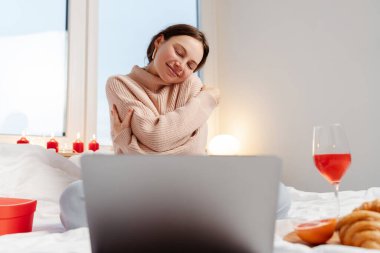 Güzel, gülümseyen bir kadının portresi. Yatakta oturuyor, kendine sarılıyor, dizüstü bilgisayar kullanıyor, video çekiyor, video kaydediyor. Çevrimiçi randevu kavramı