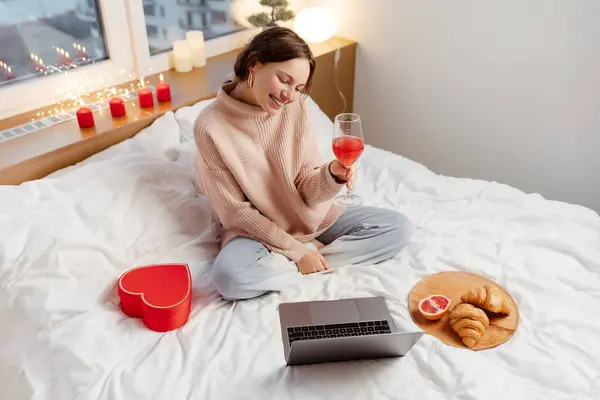 笑顔の若い女性は ラップトップコンピュータを使用してワインのグラスを保持し ボーイフレンドのお祝いバレンタインデーとオンライン通信自宅でろうそくでベッドに座っています オンラインデートコンセプト — ストック写真