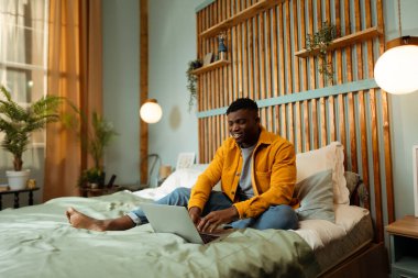 Yakışıklı, Afro-Amerikalı, hippi, renkli kıyafetler giyen, rahat bir yatakta dizüstü bilgisayar kullanan, uzaktan kumandalı bir yazar. Çevrimiçi eğitim kavramı