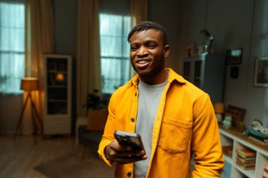 Kendine güvenen, şık sarı tişörtlü, cep telefonlu, evinde duran kameraya bakan Afro-Amerikan bir adamın portresi. Çevrimiçi teknoloji kavramı, sosyal medya