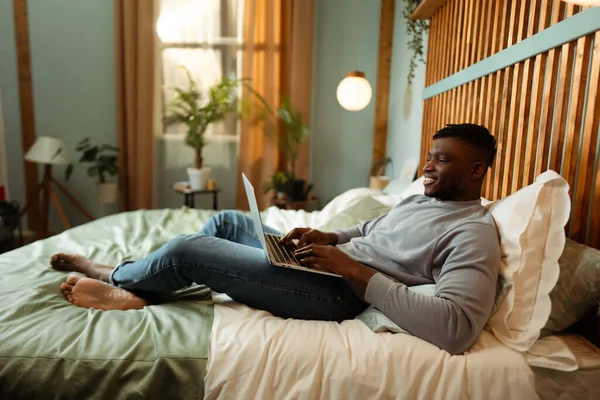 Portrait of positive happy African American man, IT specialist using laptop, lying on bed at home, relaxing, copywriter typing. Concept of remote job, online technology