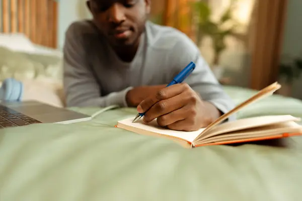 Portrait of attractive African American student lying on bed learning language, taking notes, selective focus on hand, holding pen, at home. Concept of online education, remote job