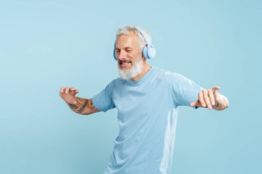 Kulaklıkla müzik dinleyen, mavi arka planda dans eden, olgun sakallı bir adamın portresi. Teknoloji konsepti, eğlence