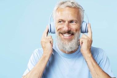 Yakışıklı, olgun, gri saçlı bir adamın portresi, kulaklıkla müzik dinleyen bir hipster, yakından bakıyor, mavi arka planda tek başına duruyor, kopyalama alanı. Teknoloji konsepti