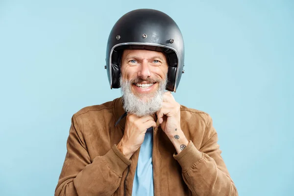 黒い保護ヘルメットを身に着けているハンサムな灰色の髪の男性バイカーの肖像画 青い背景に隔離されたカメラを見ている茶色のジャケット ホビーコンセプト — ストック写真