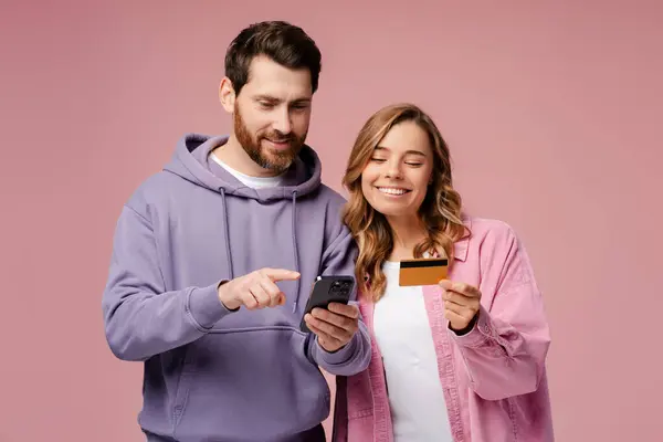 Caucasian happy smiling couple holding plastic credit card and mobile phone, isolated on pink background. Online shopping. Internet banking. Cashless payment. Paying bills. Booking and food ordering