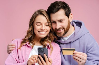 Gülümseyen bir çiftin portresi, pembe arka planda yalnız duran çekici erkek ve kadın, cep telefonu, mobil uygulama, kredi kartı, ödeme. Çevrimiçi alışveriş kavramı