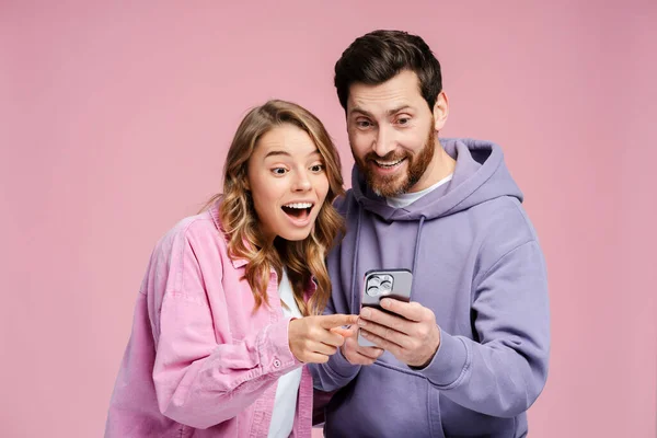 Portrait of attractive, excited couple wearing stylish colorful clothes, holding mobile phone, using mobile app, ordering standing isolated on pink background. Online shopping concept
