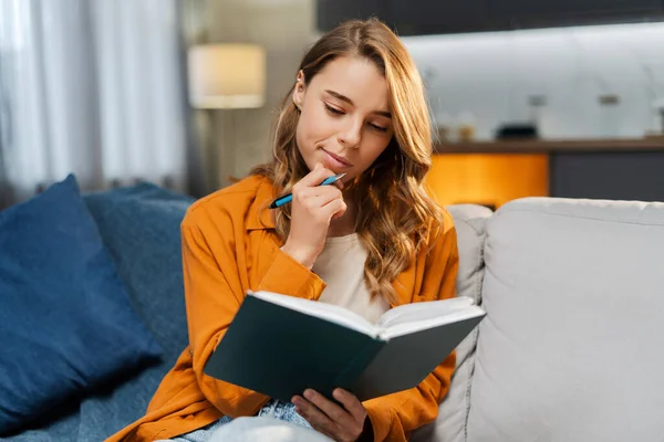 Pensive portrait of beautiful young woman, writer holding notebook and pen, taking notes while sitting on comfortable sofa at home. Concept of remote job, project planning