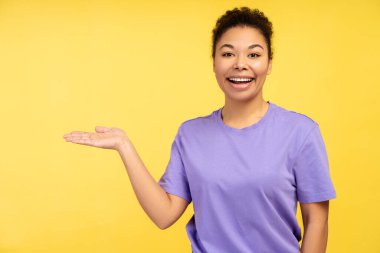 Gülümseyen, mor tişört giyen, fotokopi alanında el ele tutuşan, sarı arka planda izole edilmiş kameraya bakan güzel Afrikalı Amerikalı kadın portresi. Reklam kavramı